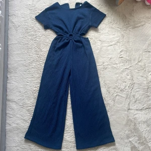 Mörkblå  flickdress med ringdetalj - Snygg mörkblå byxdress i ribbat tyg med kort ärm och breda ben. Midjan har en cool ringdetalj som ger en unik look. Tyget är mjukt och har struktur, perfekt för dig som gillar bekväma och trendiga plagg. Storlek 128  ålder 5-7år oanvänd 