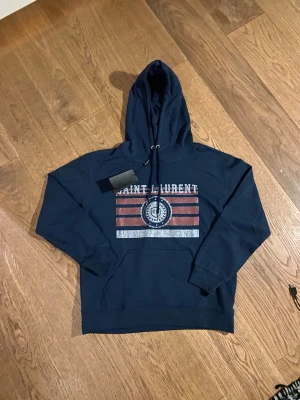 Mörkblå hoodie - Snygg mörkblå hoodie  med tryck i vitt och rött framtill. Tröjan har huva med snörning och en stor magficka. Materialet är mjuk bomull och passformen är normal. Perfekt för dig som gillar streetwear och vill ha en cool look.