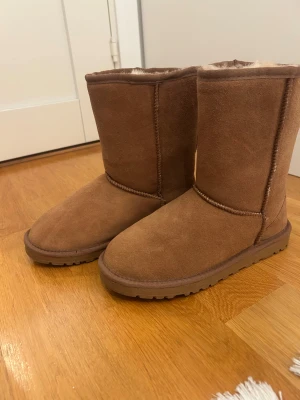 Bruna  höga boots i mocka - Säljer ett par klassiska högaboots i brun mocka med mjukt och fluffigt fårskinnsfoder. Skorna har rund tå, platt sula och är perfekta för kalla dagar.Grov yttersula för extra grepp. Supermysiga och tidlösa boots!