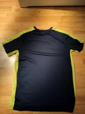 Nike svart t-shirt med gula detaljer - Säljer en sportig svart t-shirt från Nike med gula paneler längs sidorna och axlarna. T-shirten har korta ärmar och är tillverkad i ett lätt och ventilerande polyestermaterial, perfekt för träning. Klassisk Nike-logga på bröstet.