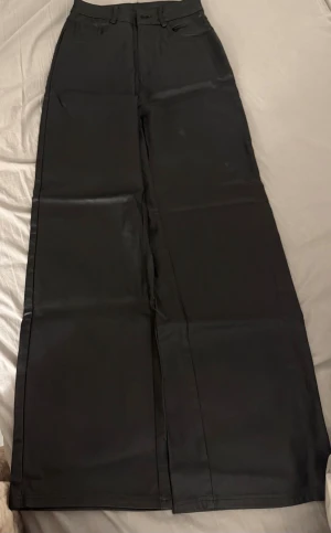 Svarta skinn byxor med hög midja. - Snygga svarta byxor med riktigt breda ben och hög midja. De har klassiska fickor både fram och bak samt bälteshällor. Materialet ser ut att vara slätt och lite glansigt, vilket ger en cool vibe. Perfekta för dig som gillar en trendig och avslappnad stil.