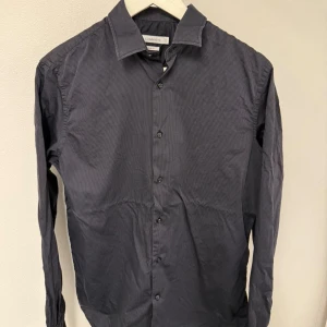 Mörkgrå skjorta J.Lindeberg - Snygg mörkgrå skjorta från J.Lindeberg i slim fit-modell. Skjortan har diskreta ränder, klassisk krage och knappar framtill. Långärmad och tillverkad i mjuk bomull som känns skön mot huden. Perfekt för dig som gillar stilrena och moderna plagg.