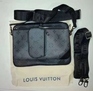 Louis Vuitton svart axelväska i skinn - Snygg svart axelväska från Louis Vuitton med klassiskt monogrammönster. Väskan har justerbar axelrem med logotyp, flera fack med dragkedja och en avtagbar mindre ficka framtill. Tillverkad i skinn med mörka detaljer och stilren design.