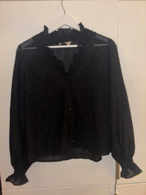 Svart glittrig blus från Åhléns  - Hejj, Säljer en svart glittrig blus från Åhléns i storlek 34. Blusen har v-ringad hals med volangkant, knappar framtill och långa ärmar som blir jätte fin nu till nyår💗🎀