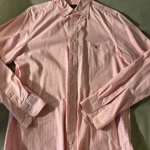 GANT skjorta rosa - Snygg rosa och vit rutig skjorta från GANT med klassisk krage och bröstficka med broderad logga. Skjortan har långa ärmar, knappar framtill och är gjord i mjuk bomull. Perfekt för dig som gillar preppy stil och vill sticka ut med färg.