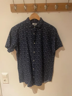 Blommig blå skjorta Jack & Jones M - Snygg kortärmad skjorta från Jack & Jones Premium i slim fit. Skjortan är mörkblå med små blommor i vitt och ljuslila, har klassisk krage och knappar framtill. Perfekt för dig som gillar stilrena detaljer och mönster.