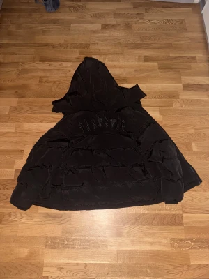 Svart pufferjacka från Trapstar - Svart pufferjacka från Trapstar med broderad logga på ryggen och avtagbar huva. Jackan har dragkedja framtill, två sidofickor och en clean look som passar streetwear. Materialet är syntet och jackan är perfekt för kalla dagar.