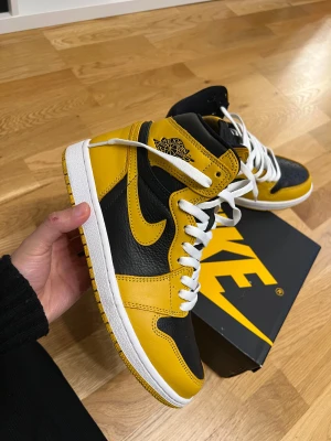 Nike Air Jordan 1 svart/gul sneakers - Nike Air Jordan 1 sneakers i svart och gul med vita snören. Klassisk high-top siluett med rund tå och platt sula. Ovandel i skinn med kontrasterande paneler och Air Jordan-logga på sidan. Snygg box med matchande färger ingår.