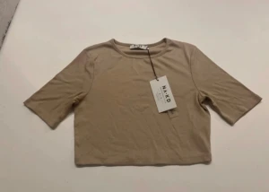 Beige croppad topp från NA-KD - Säljer en beige croppad topp från NA-KD med rund hals och halvkort ärm. Enkel och stilren design i mjukt bomullsmaterial, perfekt för att matcha med jeans eller kjol. Toppens passform är normal och den har en fräsch, modern look.