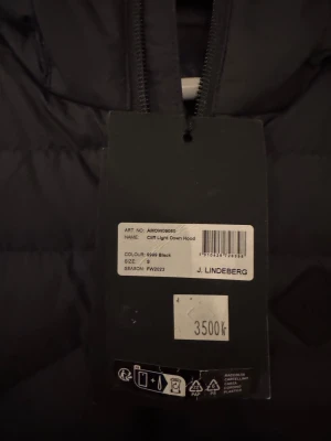 Svart dunjacka från J.Lindeberg - Svart dunjacka från J.Lindeberg med huva och snygga quiltade detaljer. Jackan har dragkedja framtill och logotyp på huvan. Perfekt för kalla dagar och har en modern, clean look som funkar till streetwear. Modellen heter Cliff Light Down Hood.