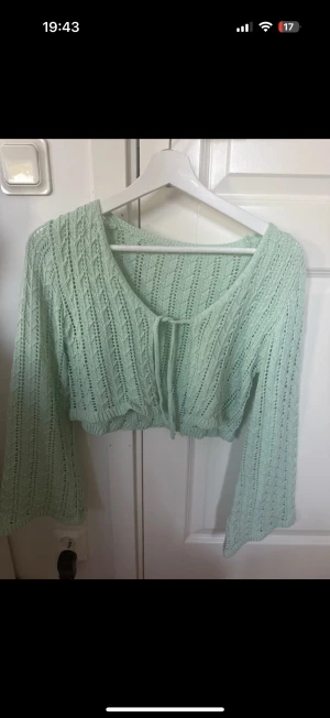 Mintgrön stickad bolerotopp - Säljer en mintgrön bolerotopp i stickat material med långärmade och croppad passform. Toppen har ett snyggt hålmönster och knyt framtill. Perfekt för att ge din outfit en fräsch och trendig touch.