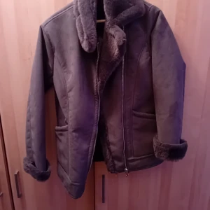 Mörkgrå pälsjacka med dragkedja - Säljer en mörkgrå pälsjacka med fluffig krage och muddar. Jackan har två fickor framtill och stängs med en rejäl dragkedja. Perfekt för kalla dagar när du vill vara både varm och snygg. Jackan har en rak passform och är helfodrad med mjuk päls på insidan.