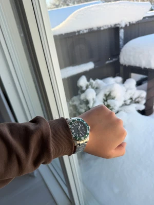 Silverfärgad klocka från Thorsen - Stilren klocka från Thorsen med rund svart urtavla och gröna detaljer på ringen. Armbandet är i silverfärgat metall och har en klassisk länkdesign. Klockan har tydliga siffror, datumvisning och en robust känsla som passar dig som gillar sportig stil.