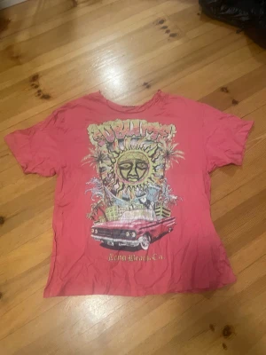 Rosa Sublime offshoulder med tryck - Säljer en rosa offshoulder från Sublime med färgstarkt tryck framtill som visar en bil, palmer och texten 'Long Beach, CA'. T-shirten har korta ärmar och en klassisk rund hals. Perfekt för dig som gillar bandmerch och streetstyle.