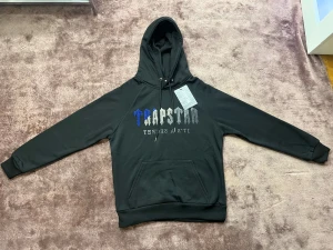 Trapstar svart hoodie med broderad logga - Svart hoodie från Trapstar i storlek S med broderad logga i blått och grått på bröstet. Tröjan har huva med snörning, magficka och ribbade muddar. Materialet är mjukt och känns som bomull och polyester. Perfekt för dig som gillar streetwear och vill sticka ut.
