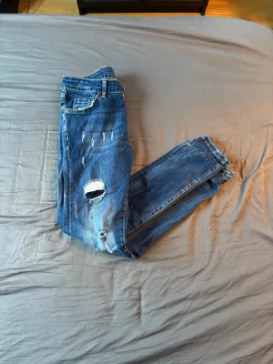 Dsquared2 blå slitna jeans - Blå jeans från Dsquared2 med coola slitningar, hål och färgstänk. Klassisk femficksmodell med patch bak på linningen. Jeansen har slim passform och är tillverkade i denim med snygg tvätt och detaljer som ger en edgy look.