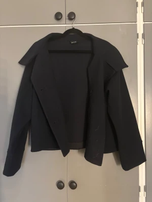 Svart skepparkavaj från Gina Tricot XS - Säljer en svart skepparkavaj från Gina Tricot i storlek XS. Jackan har klassisk krage, dubbelknäppning med svarta knappar och är tillverkad i ett ull-liknande material. Modellen är kortare och har långa ärmar, perfekt för dig som gillar stilrena och tidlösa plagg.