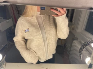 Beige stickad kofta med dragkedja - Mysig beige stickad kofta från Paul Smith med dragkedja framtill och ett broderat hundmärke på bröstet. Koftan har långärmade ärmar och ribbade muddar vid ärmslut och nederkant. Perfekt för att hålla sig varm och ändå se chill ut.