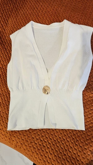 Vit stickad väst med v-ringning - Vit stickad väst med bred resår i midjan och djup v-ringning framtill. Västen har en stor dekorativ knapp framtill och är ärmlös med en croppad passform. Perfekt att styla över en skjorta eller t-shirt för en trendig look.