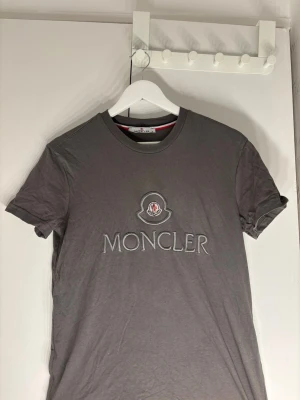 Grå Moncler t-shirt med broderad logga - Snygg grå t-shirt från Moncler i storlek S med broderad logga och emblem på bröstet. Klassisk rund hals och korta ärmar. Tillverkad i mjuk bomull som känns skön mot huden. Perfekt för dig som gillar stilrena designer med exklusiv känsla. Tveka inte på att skicka ett prisförslag.