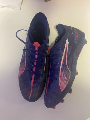 Puma Ultra blå fotbollsskor - Säljer ett par Puma Ultra fotbollsskor i blått med röda och vita detaljer. Skorna har snörning och är tillverkade i lätt syntetmaterial för bästa känsla på planen. Snygg design med mönstrad ovandel och dobbar för gräs. Perfekta för dig som vill sticka ut på matchen!