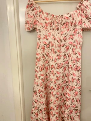 Blommig långklänning med puffärm - Superfin långklänning med blommigt mönster i rosa och brunt på ljus botten. Klänningen har puffiga korta ärmar, snörning framtill och offshoulder-design. Perfekt för sommarens alla tillfällen, med mjukt och lätt material som känns skönt mot huden.