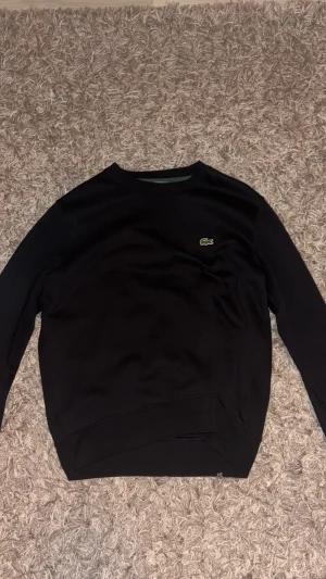 Svart Lacoste Hoodie - Svart hoodie utan luva ifrån Lacoste, storlek M. Hoodien har ett litet Lacoste märke på bröstet. Snygga svarta band med vit Lacoste-text och krokodillogga längs ärmarna. Knappt använd, har endast använts några få gånger men mest hängt i garderoben.
