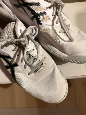 Vita Asics gympa skor  med svart logga - Säljer ett par vita Asics gympa skor med svart logga på sidan. Skorna har snörning, rund tå och platt sula. Ovandelen är i syntet och mesh för bra ventilation. Perfekta för träning eller sport. Snygg och sportig design med grå detaljer och slitstarka material. 