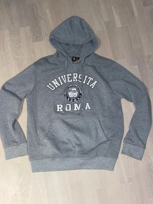  Università Roma hoodie - Grå hoodie från Chill med tryck 'Università Roma' och emblem på bröstet. Tröjan har huva med snörning, stor magficka och ribbade muddar vid ärmar och nederkant. Perfekt för dig som gillar college-vibe och bekväma plagg.