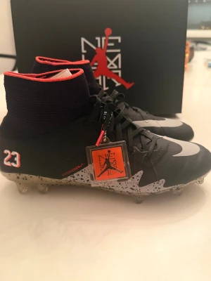 Nike Hypervenom x Jordan fotbollsskor - Nike Hypervenom x Jordan fotbollsskor i svart med röda och vita detaljer, nummer 23 broderat på sidan och Jumpman-logga på hälen. Skorna har högt skaft i stickad syntet, snörning och en platt sula med dobbar i beige och svart prickigt mönster. Snygg design för dig som gillar sport och streetstyle.