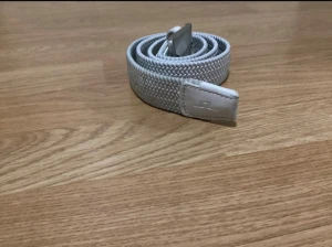 Ljusgrått flätat bälte J.Lindeberg - Snyggt ljusgrått flätat bälte från J.Lindeberg med silverfärgat spänne och detaljer i skinn. Bältet har en stilren design med diskret logga på läderdetaljen och passar perfekt till jeans eller chinos. Flätad struktur ger en cool och modern känsla.