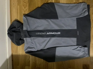 Grå vindjacka från Under Armour - Snygg grå vindjacka från Under Armour med huva och hel dragkedja. Jackan har två olika grå nyanser, logga på bröstet och stor Under Armour-tryck på ryggen. Perfekt för träning utomhus, med långärmade ärmar och lätt syntetmaterial. Kvitto finns.