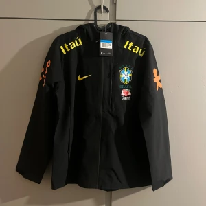 Svart Brasil Nike vindjacka med huva - Svart vindjacka från Nike med Brasil CBF-logga, gula och orangea tryck samt huva. Jackan har dragkedja framtill, långa ärmar och flera sponsormärken. Perfekt för dig som gillar fotboll och vill ha en sportig stil.