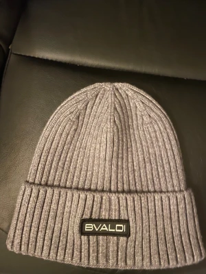 Grå ribbstickad mössa från BVALDI - Snygg grå ribbstickad mössa från BVALDI med uppvikt kant och svart logotyp framtill. Tillverkad i en mjuk blandning av viskos, nylon och polyester. Perfekt för kalla dagar och enkel att matcha med din stil.helt ny.Nypris 299.Mitt pris 149kr