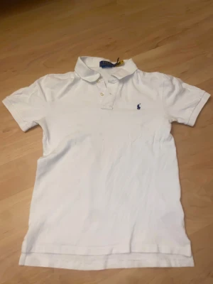 Vit slim fit piké från Polo Ralph Lauren - Vit pikétröja från Polo Ralph Lauren i slim fit med klassisk krage och korta ärmar. Tröjan har två knappar framtill och den ikoniska blå broderade loggan på bröstet. Tillverkad i mjuk bomull som är skön mot huden.