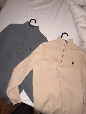Ralph Lauren Quarter Zips - Två stickade tröjor från Polo Ralph Lauren, en i beige och en i grått. Båda har half zip och klassisk broderad logga i marinblått på bröstet. Tröjorna har hög krage och långärmad passform, perfekta för att hålla värmen och se clean ut.