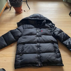 Svart pufferjacka från Polo Ralph Lauren - Svart pufferjacka från Polo Ralph Lauren med huva och röda logotypen broderad på bröstet. Använd endast förra vintern, fint skick. Finns i Umeå eller fraktas😃
