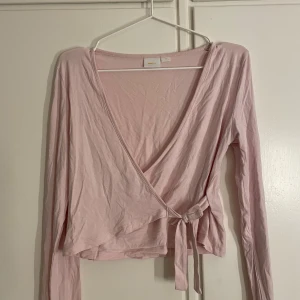 Ljusrosa omlottopp med knyt - Säljer en ljusrosa omlottopp från Gina med långa ärmar och snygg knytning i sidan. Toppen har en djup v-ringning och är gjord i mjuk bomull som känns skön mot huden. Perfekt för dig som gillar en feminin och chill stil. Storlek 170 men passar mig som bär S.