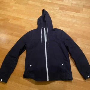 Mörkblå vindjacka med huva - Snygg mörkblå vindjacka med vit dragkedja och vita snören i huvan. Jackan har två fickor med vita knappar och är långärmad. Perfekt för blåsiga dagar och har en clean, enkel design som funkar till många stilar.