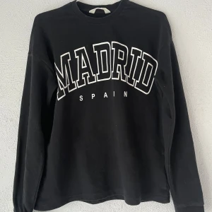 Svart sweatshirt MADRID H&M - Svart sweatshirt från H&M med stort vitt tryck 'MADRID SPAIN' på bröstet. Klassisk rund halsringning och långa ärmar. Skön och avslappnad passform, perfekt till jeans eller mjukisbyxor.