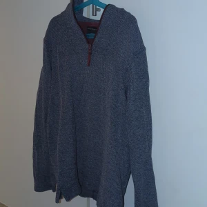 Grå stickad tröja - Säljer en grå stickad tröja från Docklands i storlek medium. Tröjan har en snygg half zip-dragkedja med vinröda detaljer vid kragen och dragkedjan. Långärmad och perfekt för kyligare dagar. Enkel och stilren design som funkar till det mesta.