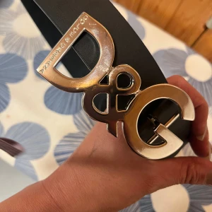 Svart D&G bälte med silverdetalj - Snyggt svart bälte från Dolce & Gabbana med stor, blank silverfärgad D&G-logga som spänne. Bältet är i skinn och har präglad text längs remmen. Perfekt för att lyfta vilken outfit som helst med en lyxig touch.