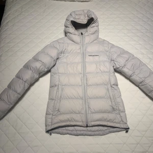 Ljusgrå dunjacka Peak Performance XS - Säljer en ljusgrå dunjacka från Peak Performance i storlek XS. Jackan har huva, quiltad design och dragkedja framtill. Perfekt för kalla dagar och har en clean look med diskret logga på bröstet. Snygg och funktionell för dig som gillar stilren vinteroutfit.