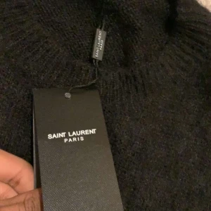 Svart Saint Laurent stickad tröja - Svart stickad tröja från Saint Laurent med vit, fluffig logotyp framtill. Tröjan har rund hals och långa ärmar, och är gjord i ett mjukt material som ger en lyxig känsla. Perfekt för dig som vill ha en statement piece med designer-vibe.