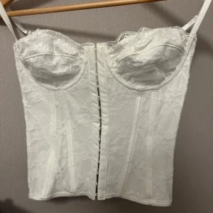 Vit spetskorsett med snörning - Säljer en vit korsett med spetsdetaljer och justerbara axelband. Framsidan har hakknäppning och baksidan har snygg snörning för extra attityd. Kuporna är formade och ger en schysst siluett. Perfekt för dig som vill ha en statement piece i garderoben.