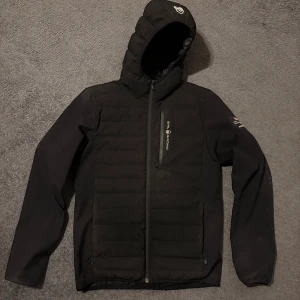 Svart pufferjacka från Sail Racing - Svart pufferjacka från Sail Racing med huva. Jackan har dragkedja, långärmade ärmar och en bröstficka med logotyp. Materialet är syntet och polyester, perfekt för kyliga dagar. Snygg och sportig look med diskreta detaljer. Har en liten defekt nere vid höger ficka och sedan så är den lite smutsig på vänster armbåge men annars i bra skick. 