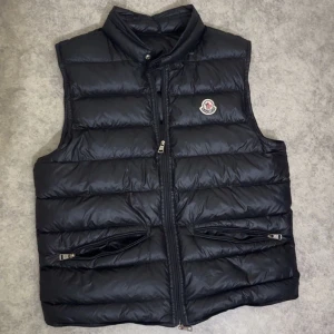 Svart dunväst från Moncler - Säljer en svart dunväst från Moncler med klassisk logga på bröstet. Västen har quiltad design, hög krage och två fickor med dragkedja framtill. Materialet är glansig polyester och dunfyllning, perfekt för kyliga dagar. Snygg och stilren look med dragkedja i mitten.storlek S men passar xs OBS! Har en jätte liten skada på ryggen man märker inte den