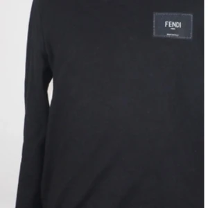 Svart långärmad tröja från Fendi - Svart långärmad tröja från Fendi med rund halsringning och diskret Fendi-logga på bröstet. Tröjan har en klassisk passform och är tillverkad i mjuk bomull, perfekt för en clean och stilren look.