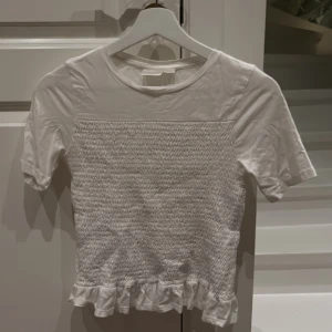 Vit croppad topp med volang från Gina Tricot - Supersöt vit croppad topp från Gina Tricot i storlek 158/164. Toppen har korta ärmar, smockad framsida och baksida samt en gullig volangkant längst ner. Perfekt för dig som gillar en feminin och trendig stil. Lite skrynklig, men kommer stryka den innan jag levererar isåfall.