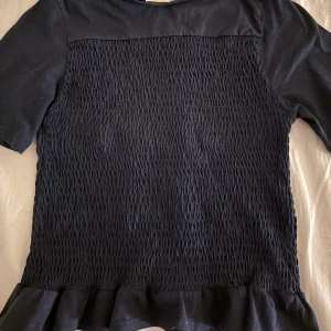 Marinblå  blus med volangkant - Säljer en marinblå blus med korta ärmar och snyggt rynkad framsida. Blusen har en volangkant längst ner som ger en extra touch. Materialet känns mjukt och lätt